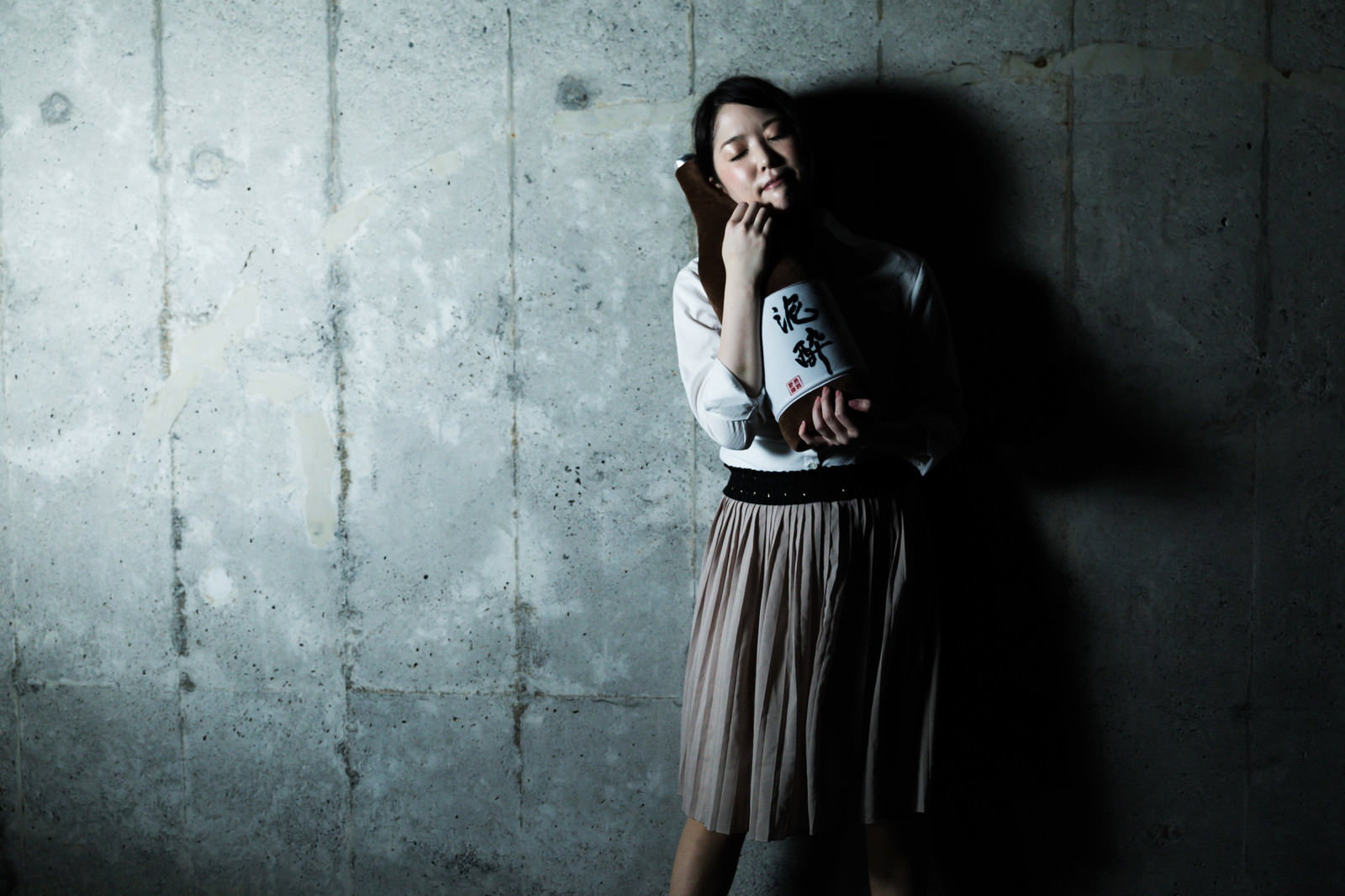 グレーのコンクリート壁にもたれ、左手を顔に当ててうつむく泥酔した女性［モデル：シマヅ］