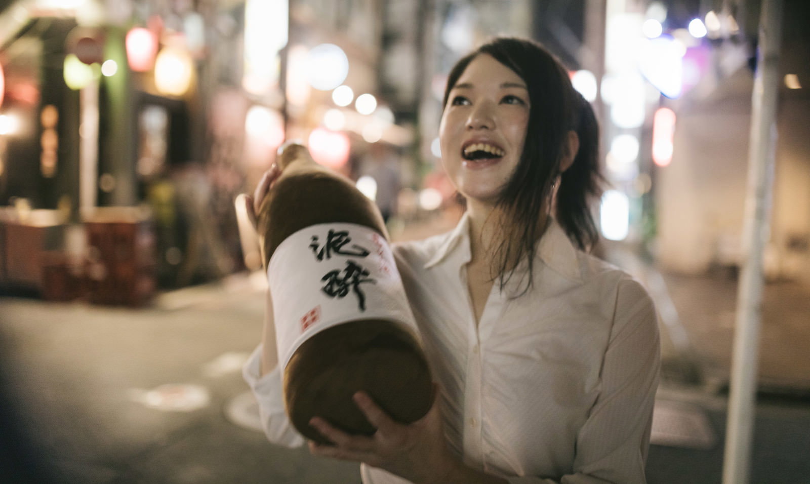 夜の街でお酒を持って笑顔を見せる酔った女性［モデル：シマヅ］