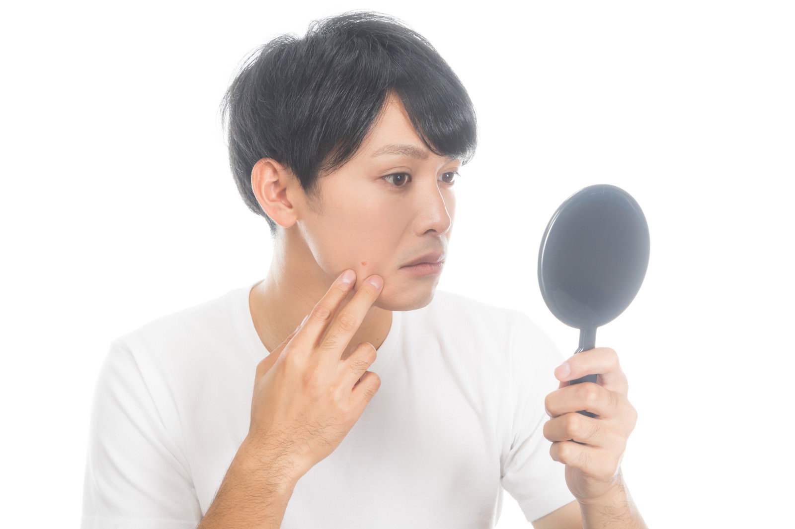 手鏡を持って頬の肌荒れをチェックしている白いシャツの男性［モデル：完伍］