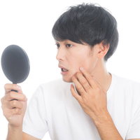 不摂生で頬に吹き出物ができた男性が手鏡でチェックの写真