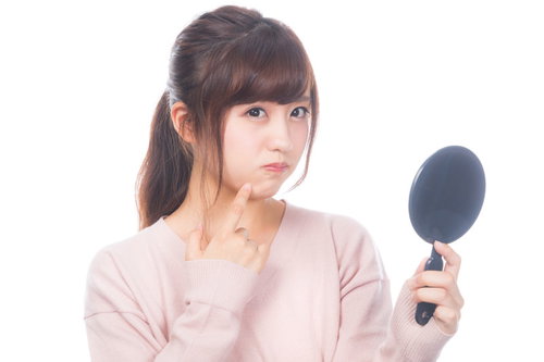 手鏡で肌のニキビをチェックして不満顔の女性