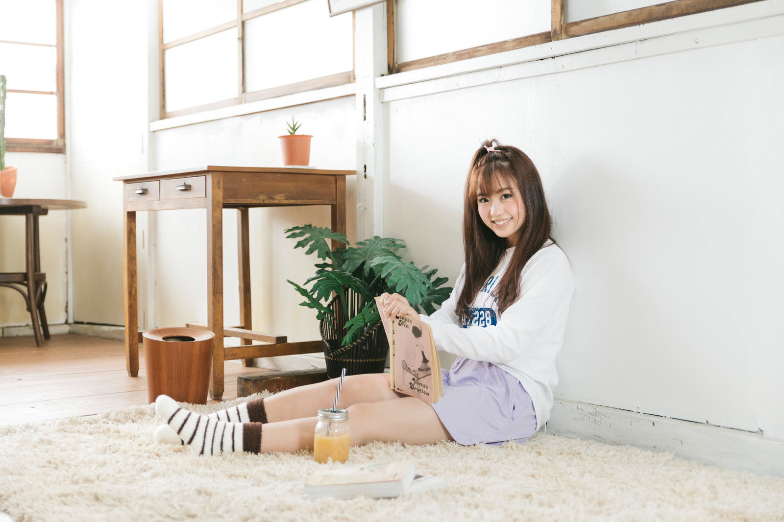 部屋でラグの上に座り、本を読みながらスムージーを飲む女性［モデル：河村友歌］
