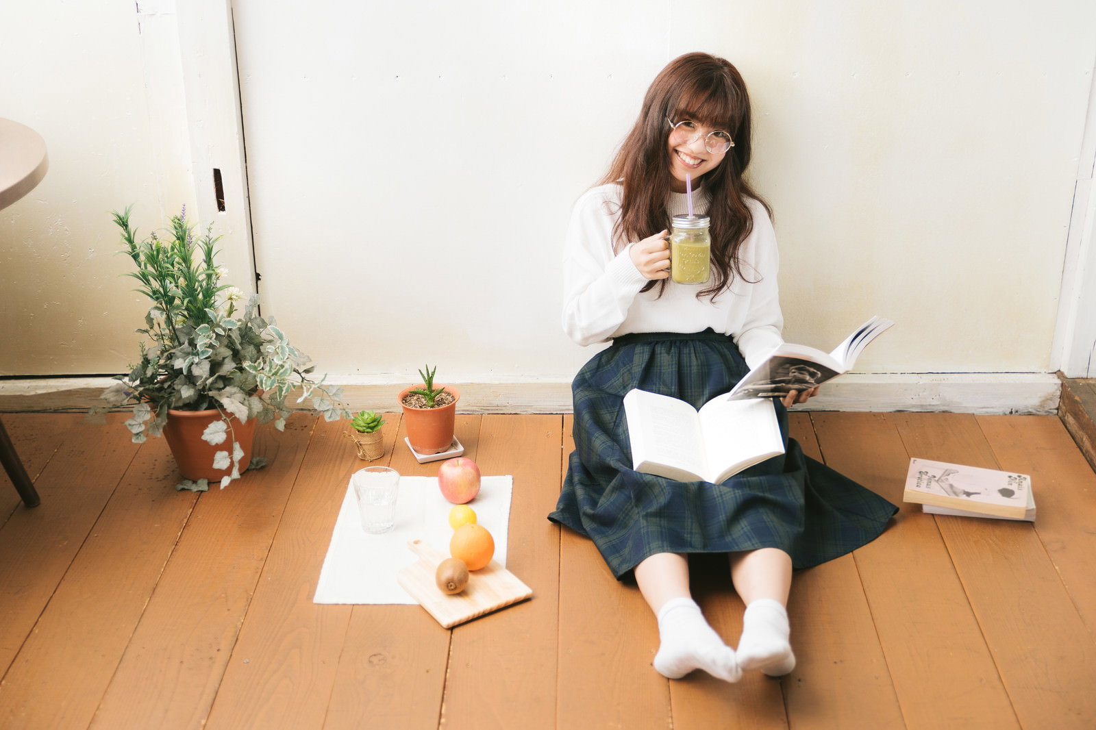グリーンスムージーを片手に床に座って本を読む笑顔の女性［モデル：河村友歌］