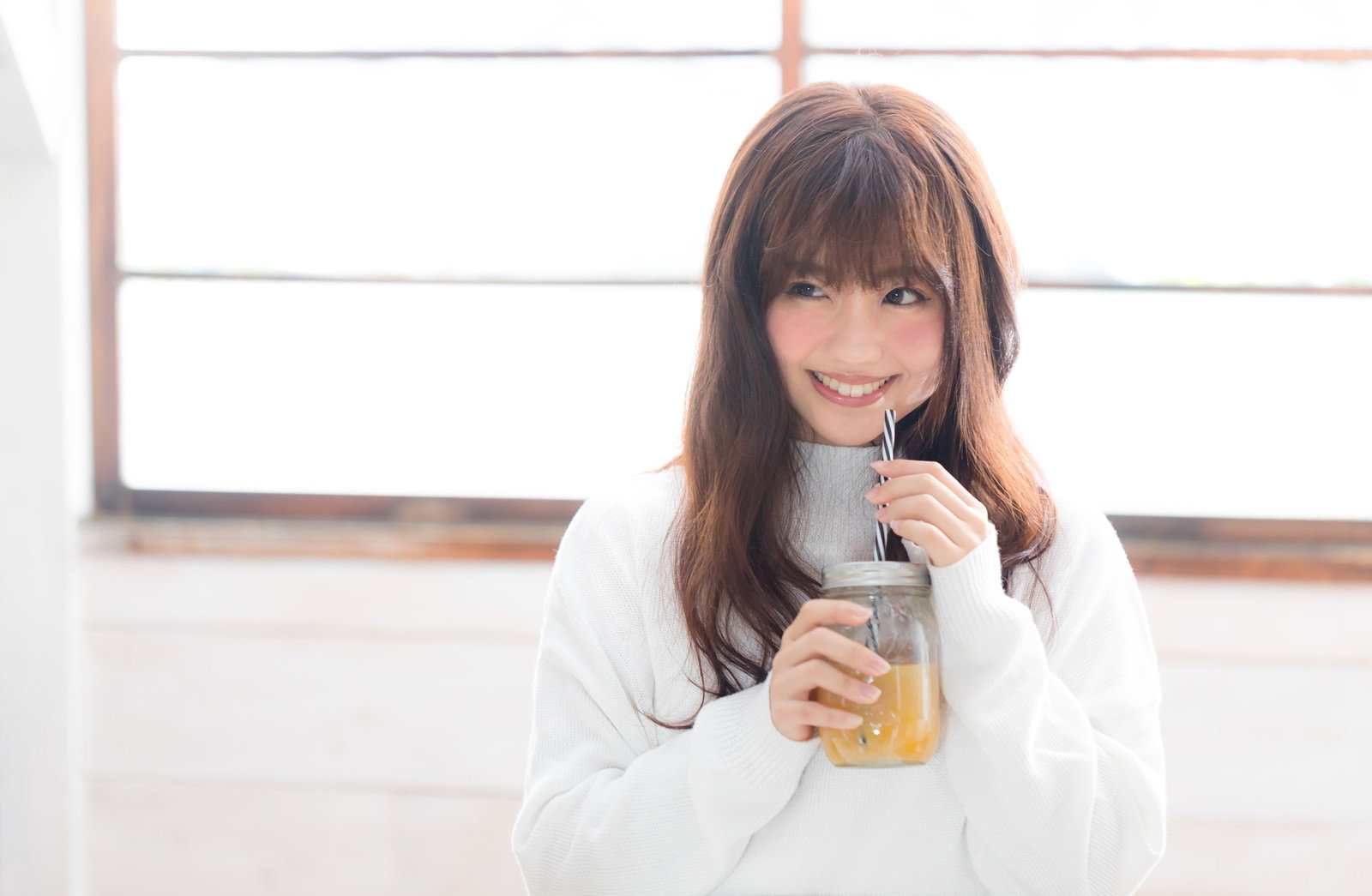白いセーター姿で瓶入りスムージーを飲んで笑う女性［モデル：河村友歌］
