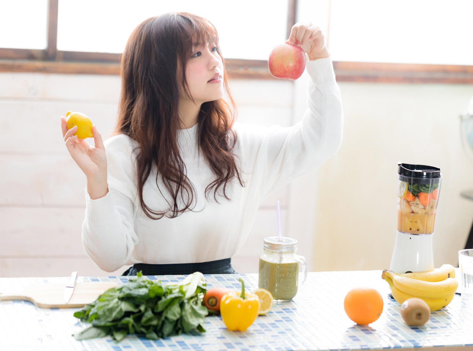 キッチンテーブルでスムージー用の果物を選ぶ女性。りんごとレモンを両手に持ち上げている［モデル：河村友歌］