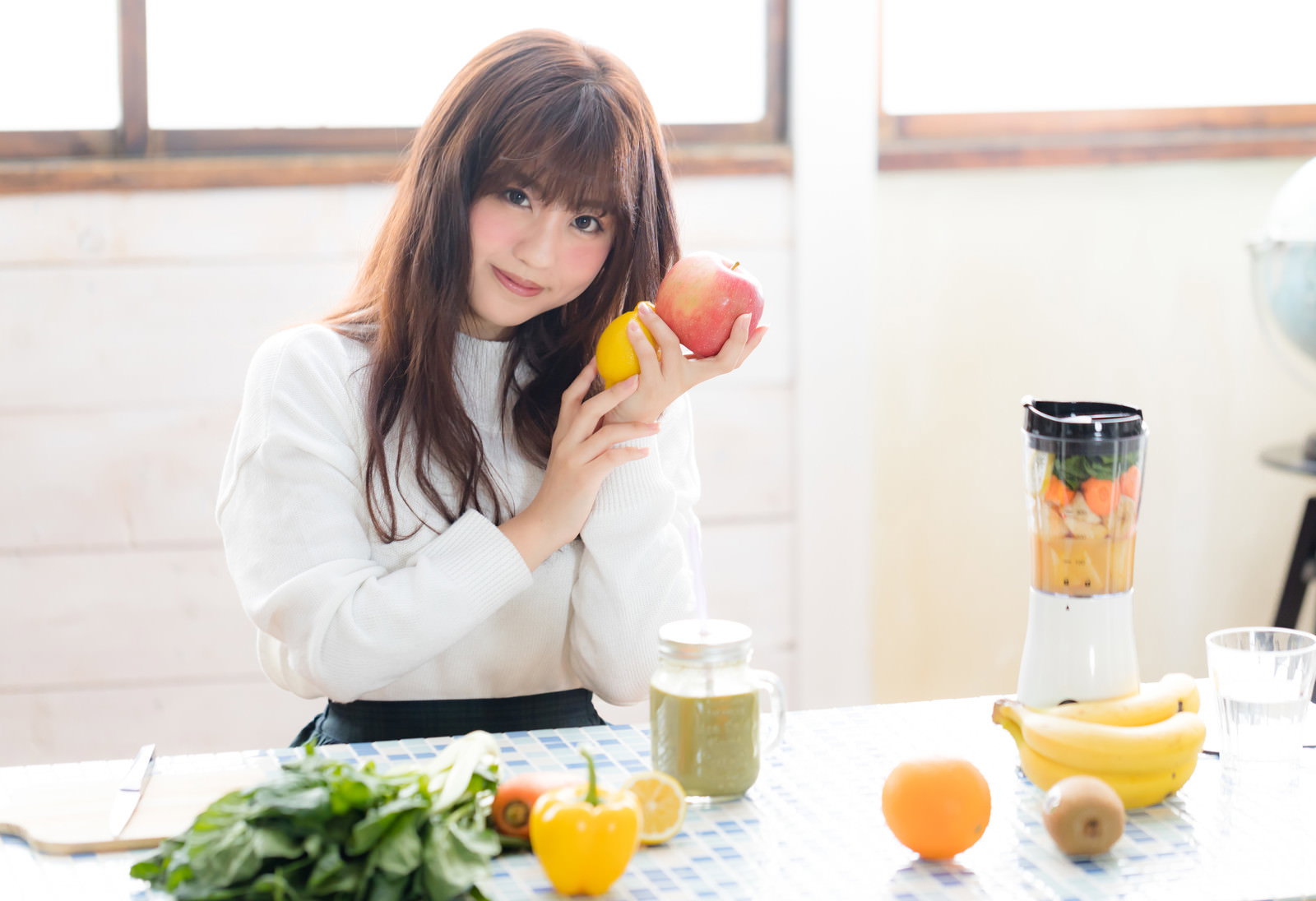 テーブルでリンゴを持って微笑む女性。ミキサーとスムージー、新鮮な果物が並ぶ［モデル：河村友歌］