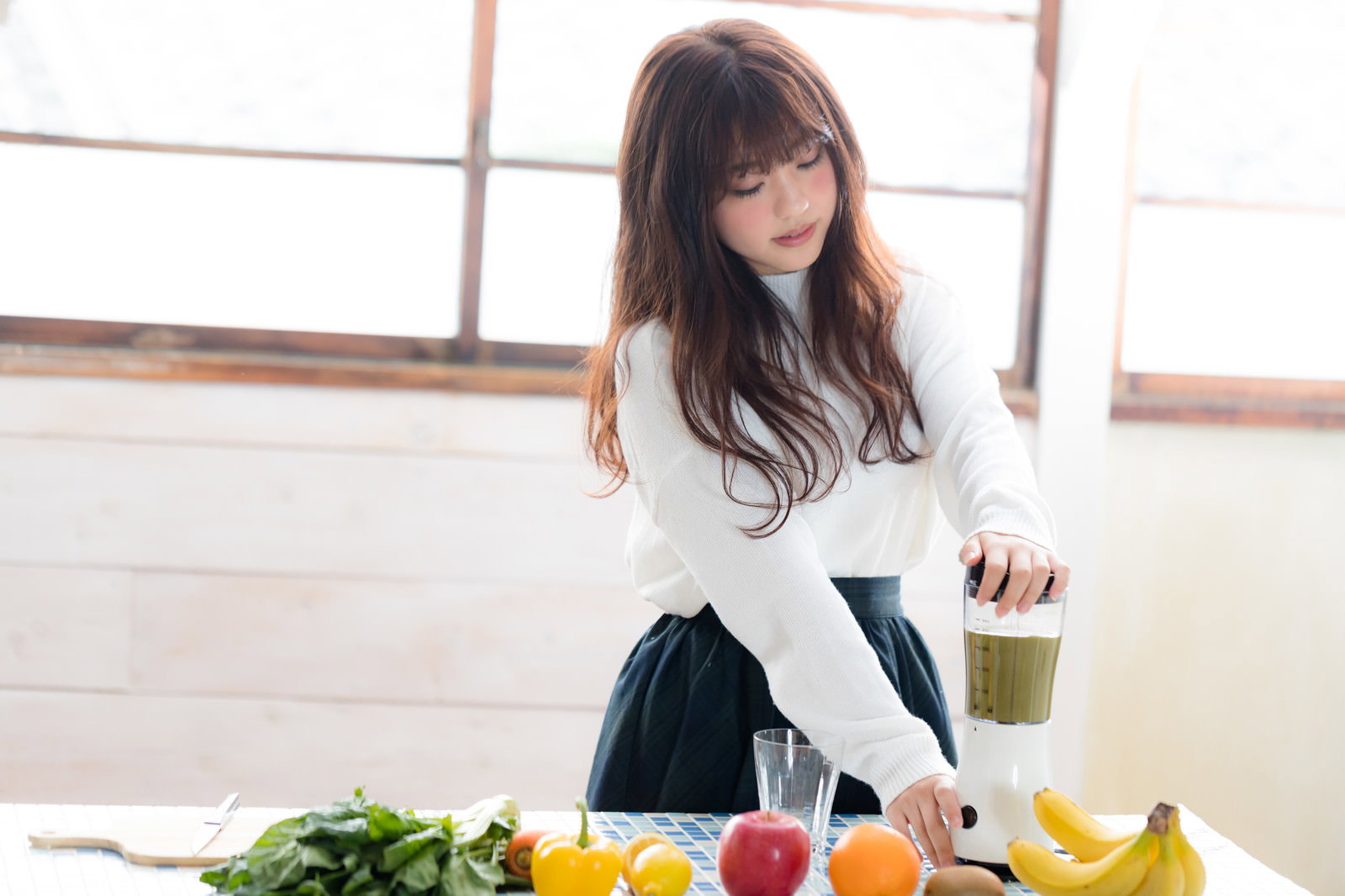 明るいキッチンでジューサーを使ってグリーンスムージーを作る白いトップスの女性［モデル：河村友歌］