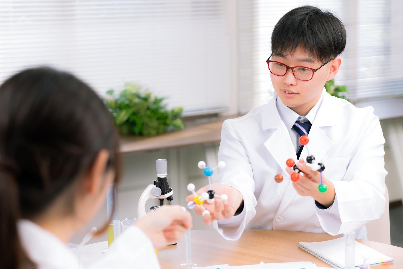 白衣を着た男性が分子モデルを持ち、同僚に説明している研究室での一場面［モデル：モデルリリース］