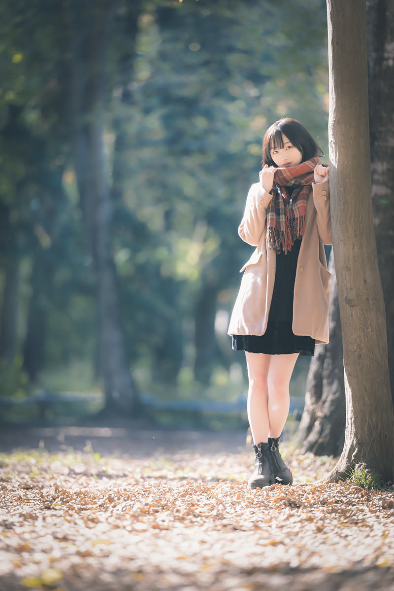 秋の公園で木にもたれかかるベージュのコートを着た女性［モデル：井隼そら］