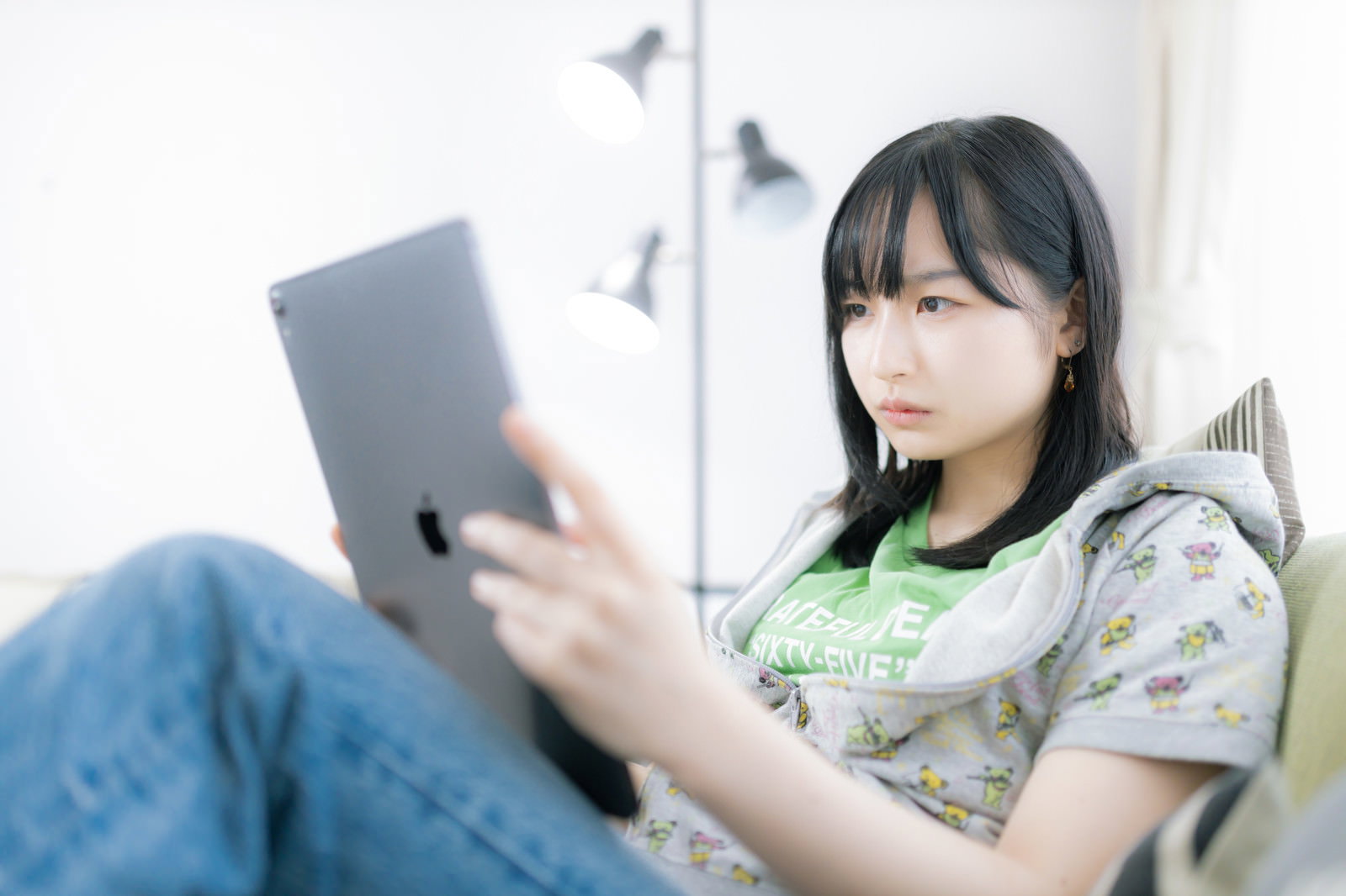 ソファに座ってタブレットを見ながら困惑した表情を見せる女性［モデル：井隼そら］