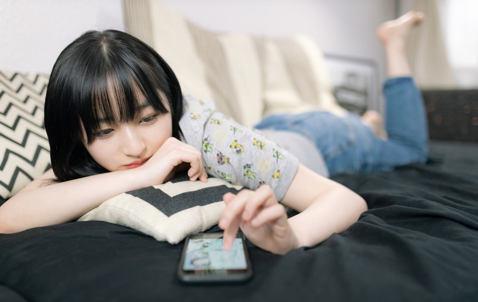ベッドでスマートフォンを見ながらくつろいでいる黒髪の女性［モデル：井隼そら］
