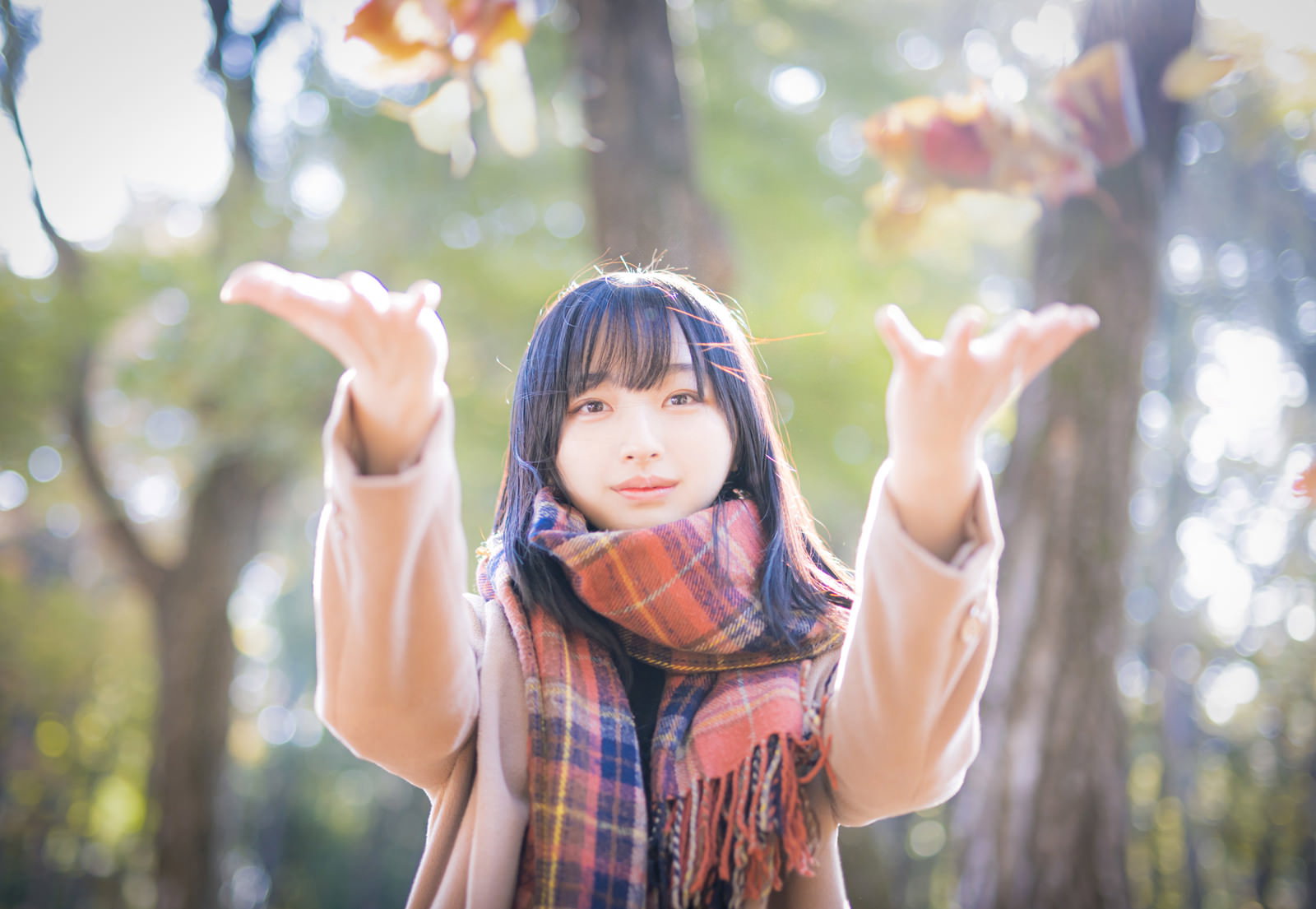 落ち葉を両手で投げ上げる茶色のマフラーを着けた女性［モデル：井隼そら］