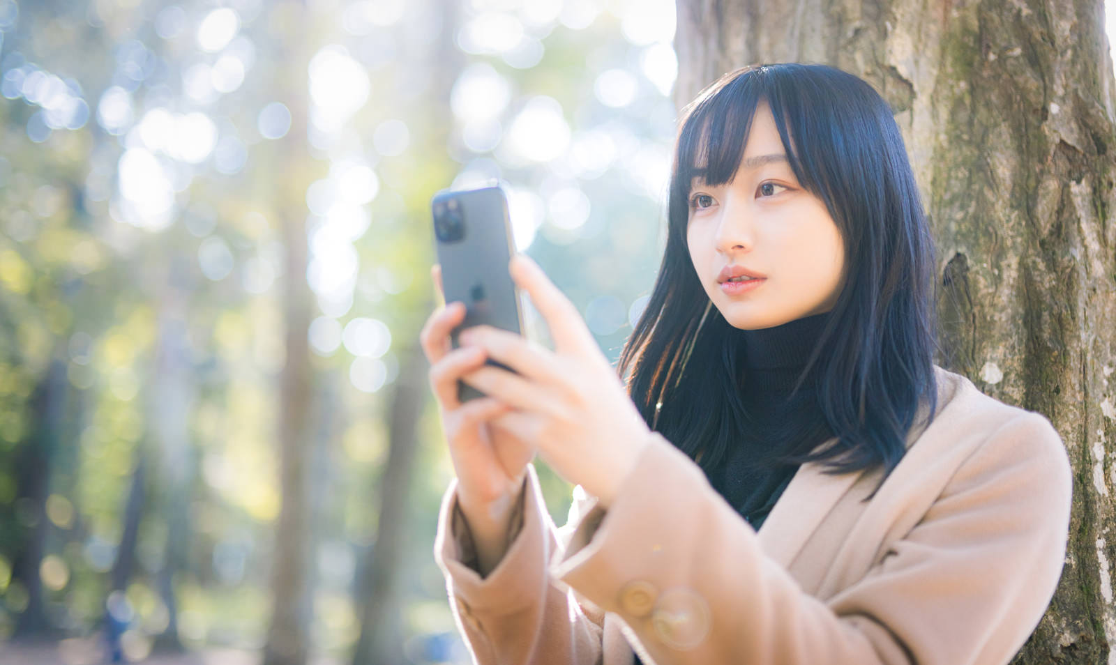 木漏れ日の中でスマートフォンを両手で持ち画面を見る女性［モデル：井隼そら］