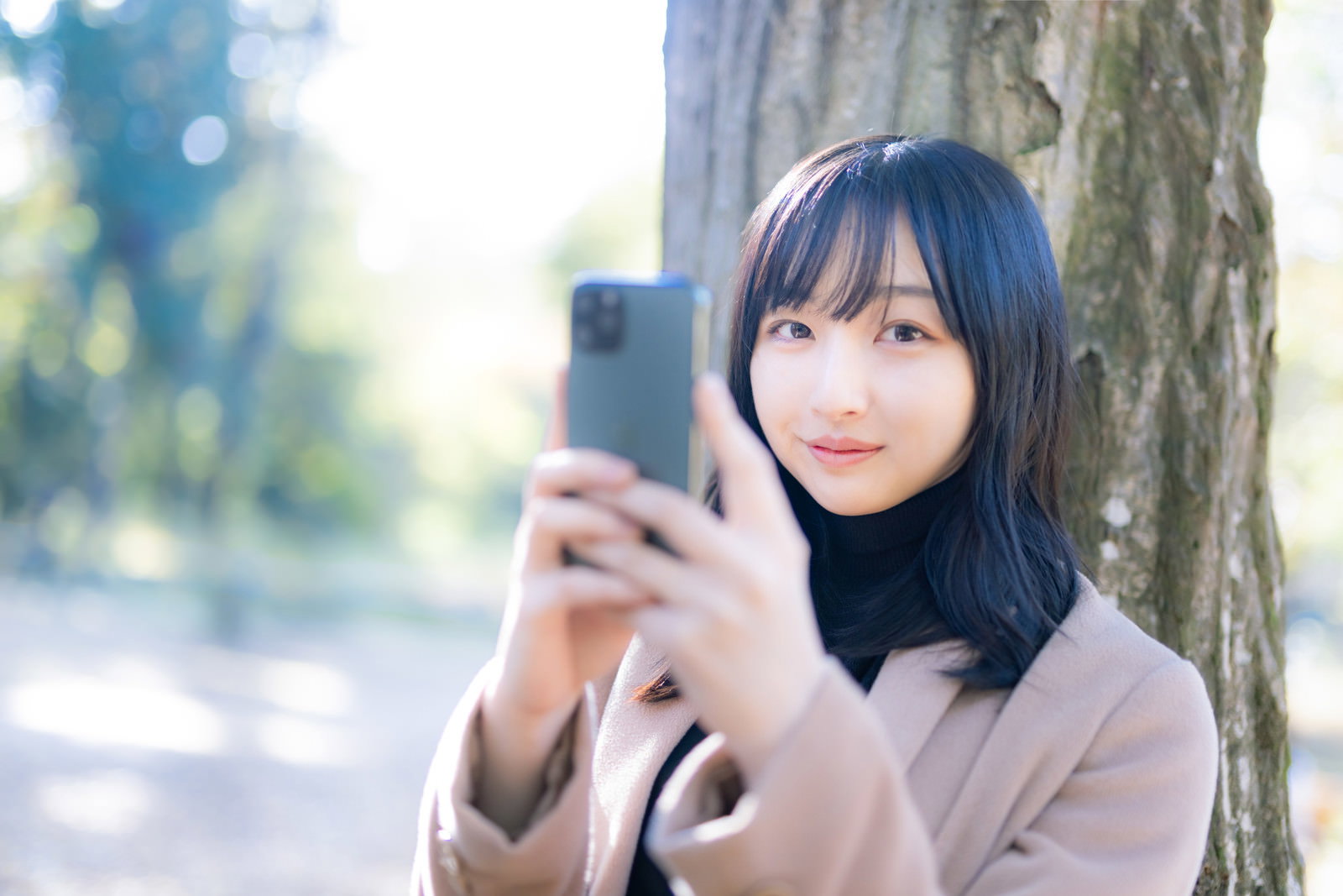 木に寄りかかりながらスマートフォンを見る女性［モデル：井隼そら］