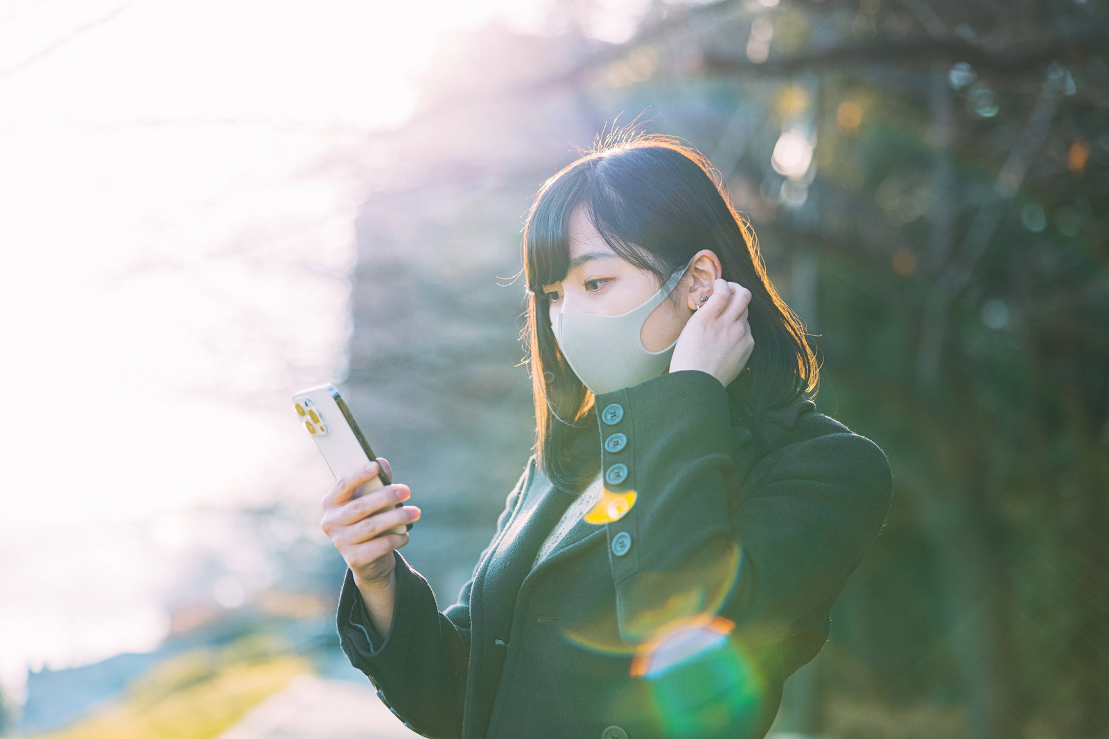 夕暮れ時にマスクを着用してスマートフォンでメッセージをチェックする女性［モデル：井隼そら］