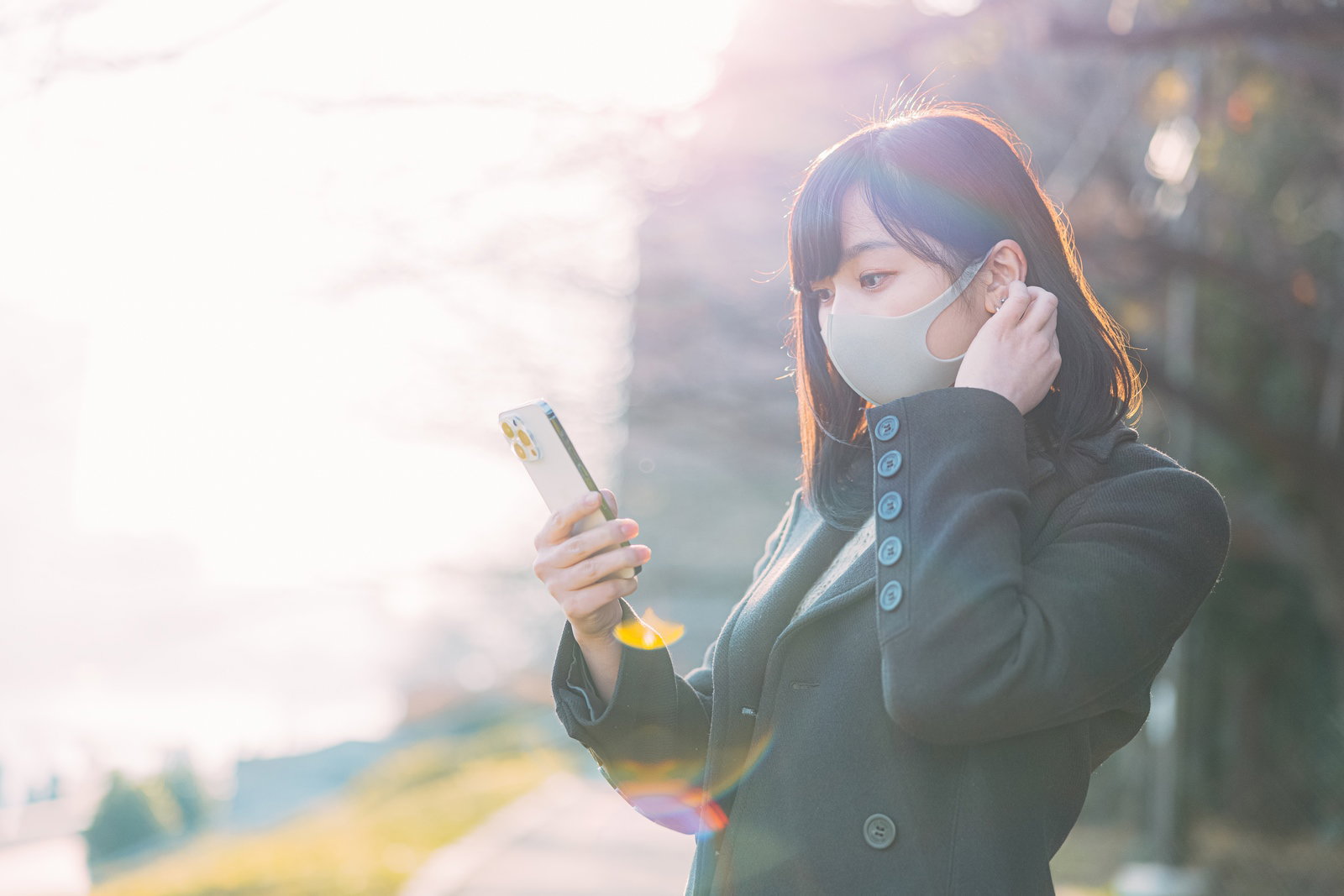 マスクを着けてスマートフォンを操作する女性が屋外で待っている［モデル：井隼そら］