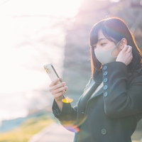 待ちぼうけでスマホを手に取るマスク姿の女性の写真