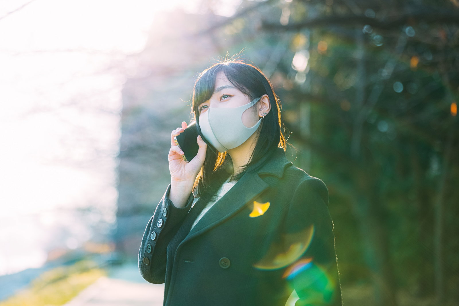 白いマスクを着用した女性が緑の屋外でスマートフォンで通話している［モデル：井隼そら］
