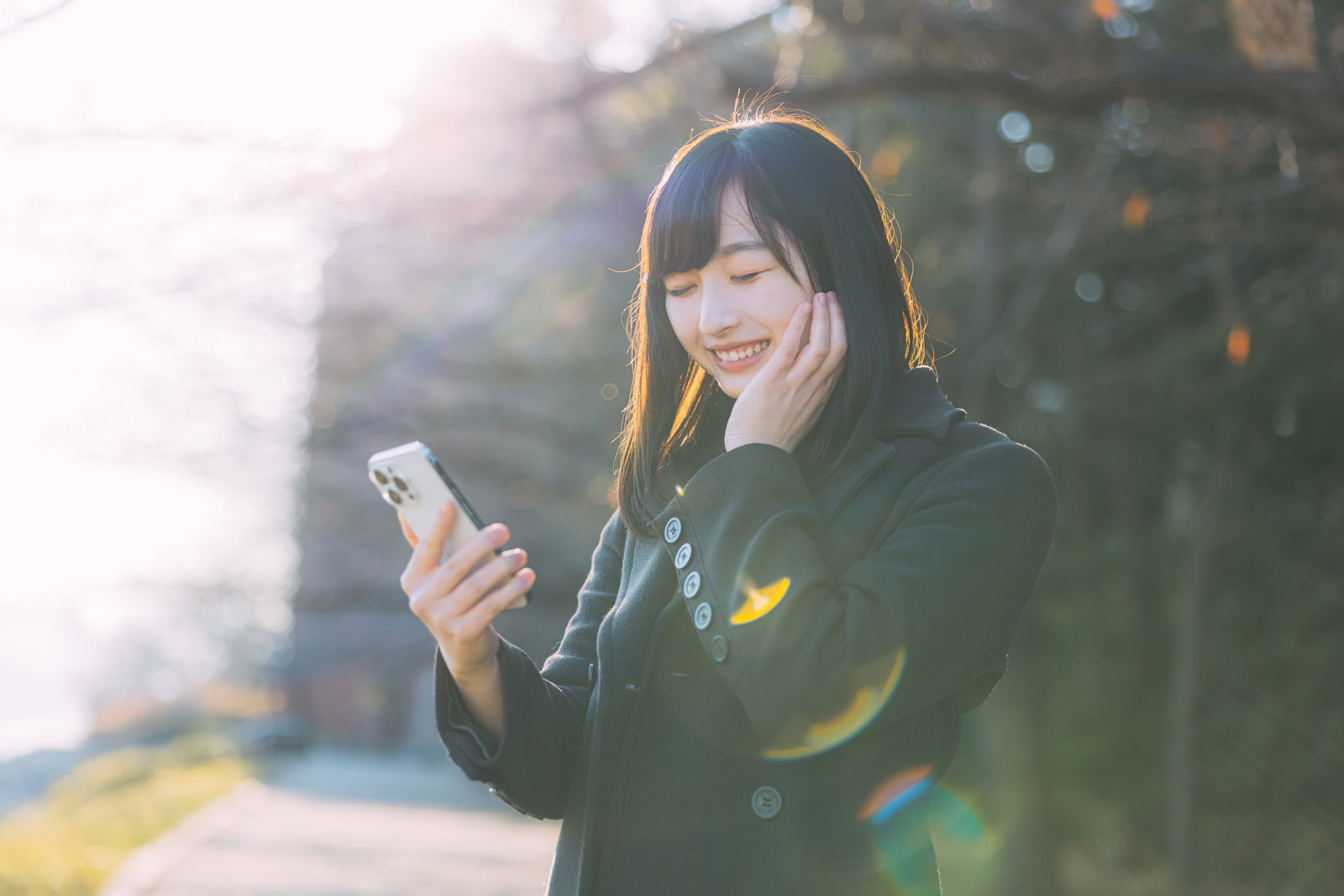 夕暮れの中でスマートフォンを見て嬉しそうに笑顔を見せる女子大生［モデル：井隼そら］