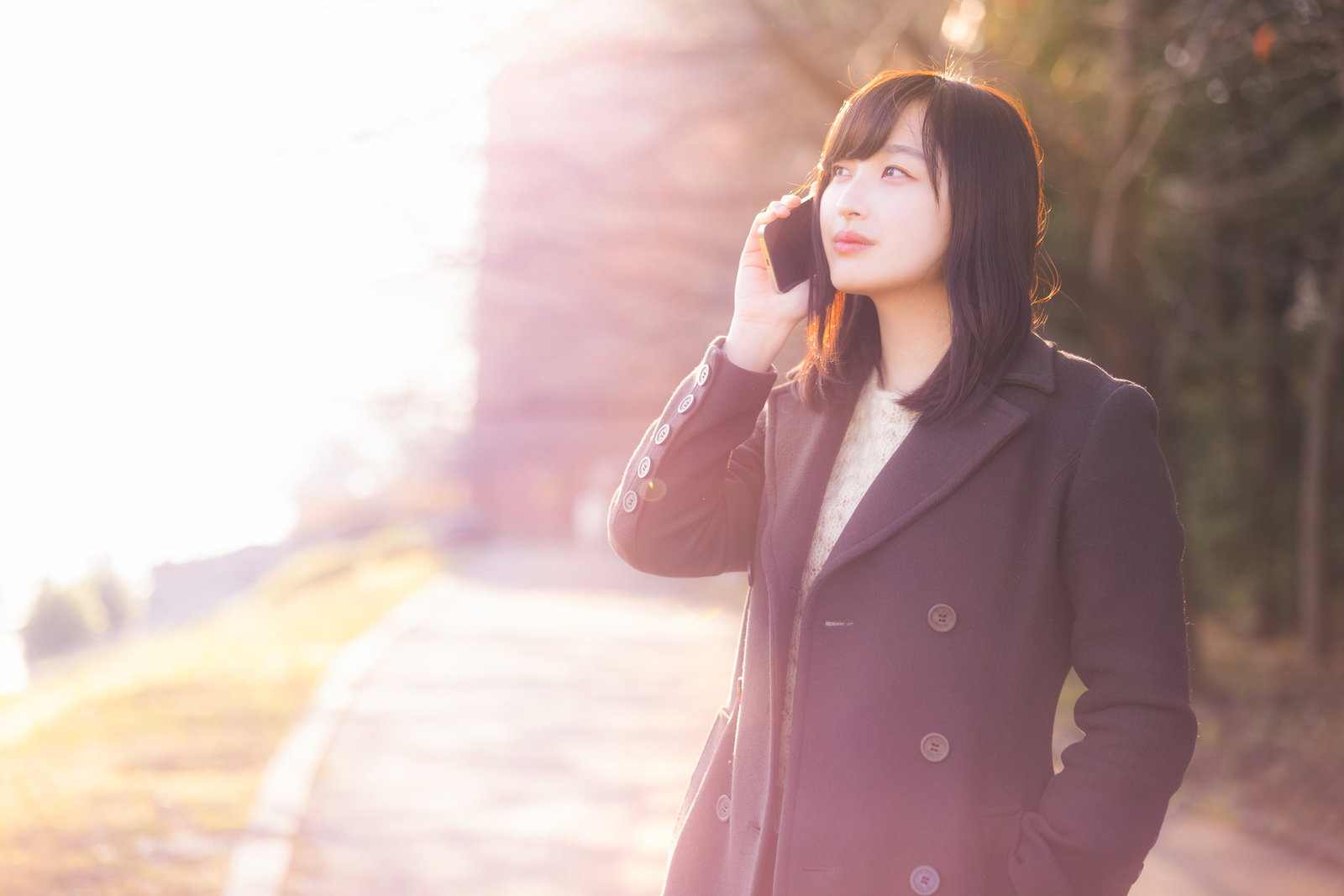 黄昏時の逆光の中で携帯電話で通話するコート姿の女性［モデル：井隼そら］