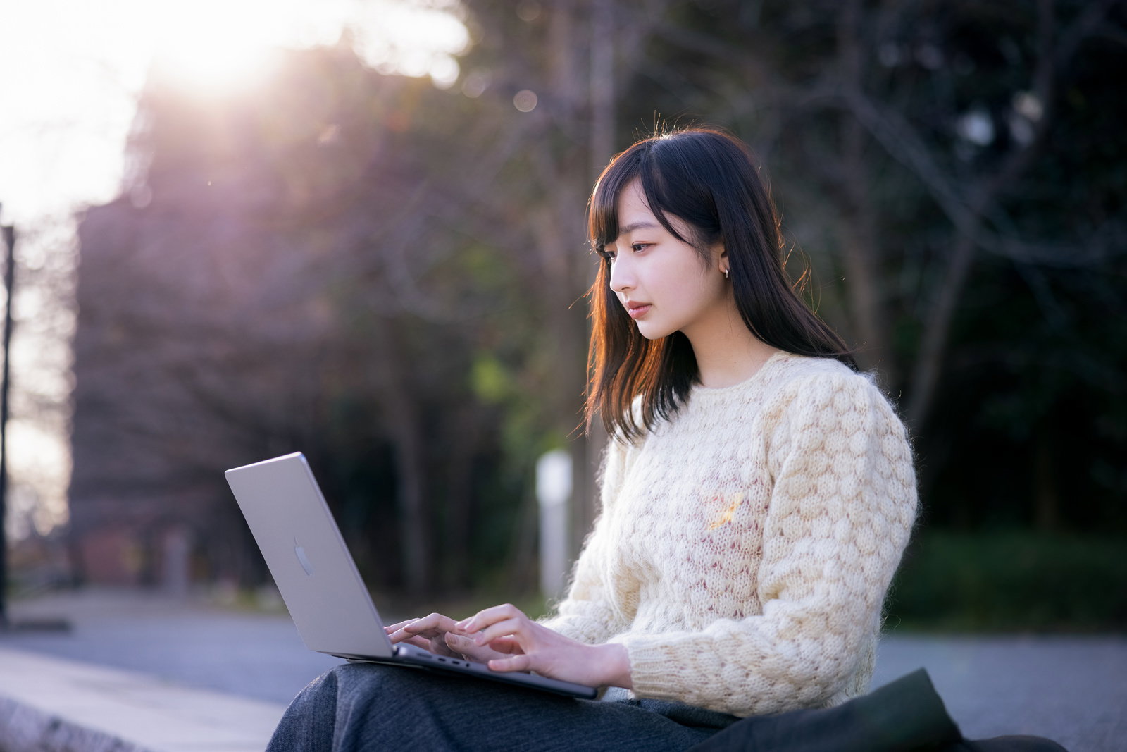 白いセーターを着た女性が屋外でノートパソコンを操作している様子［モデル：井隼そら］
