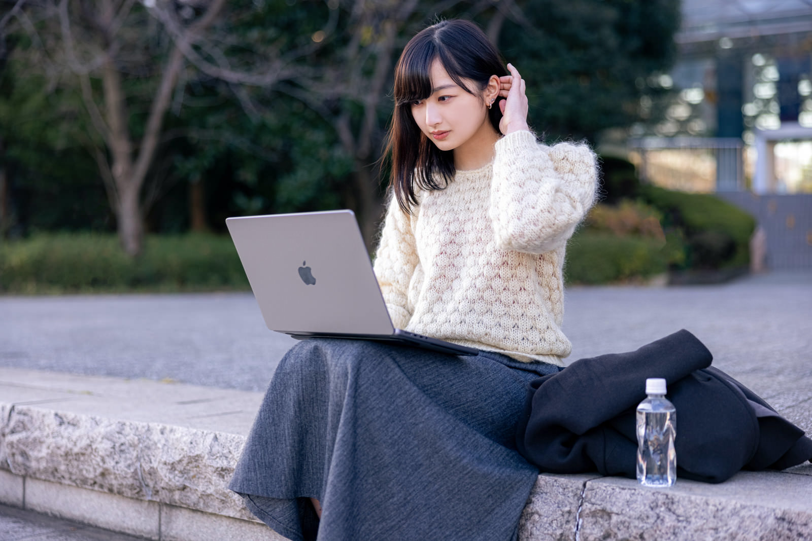 屋外のコンクリートの縁石に座り、膝の上にノートパソコンを置いて作業する女性［モデル：井隼そら］