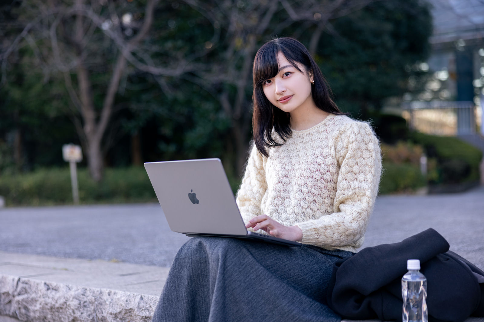 公園のベンチでMacbookを使って作業する笑顔の女性［モデル：井隼そら］