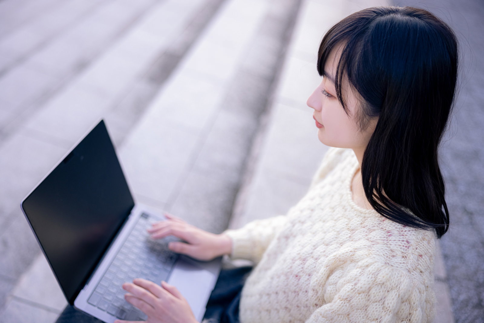 白いニットを着た女性が床に座りノートパソコンで作業している様子［モデル：井隼そら］