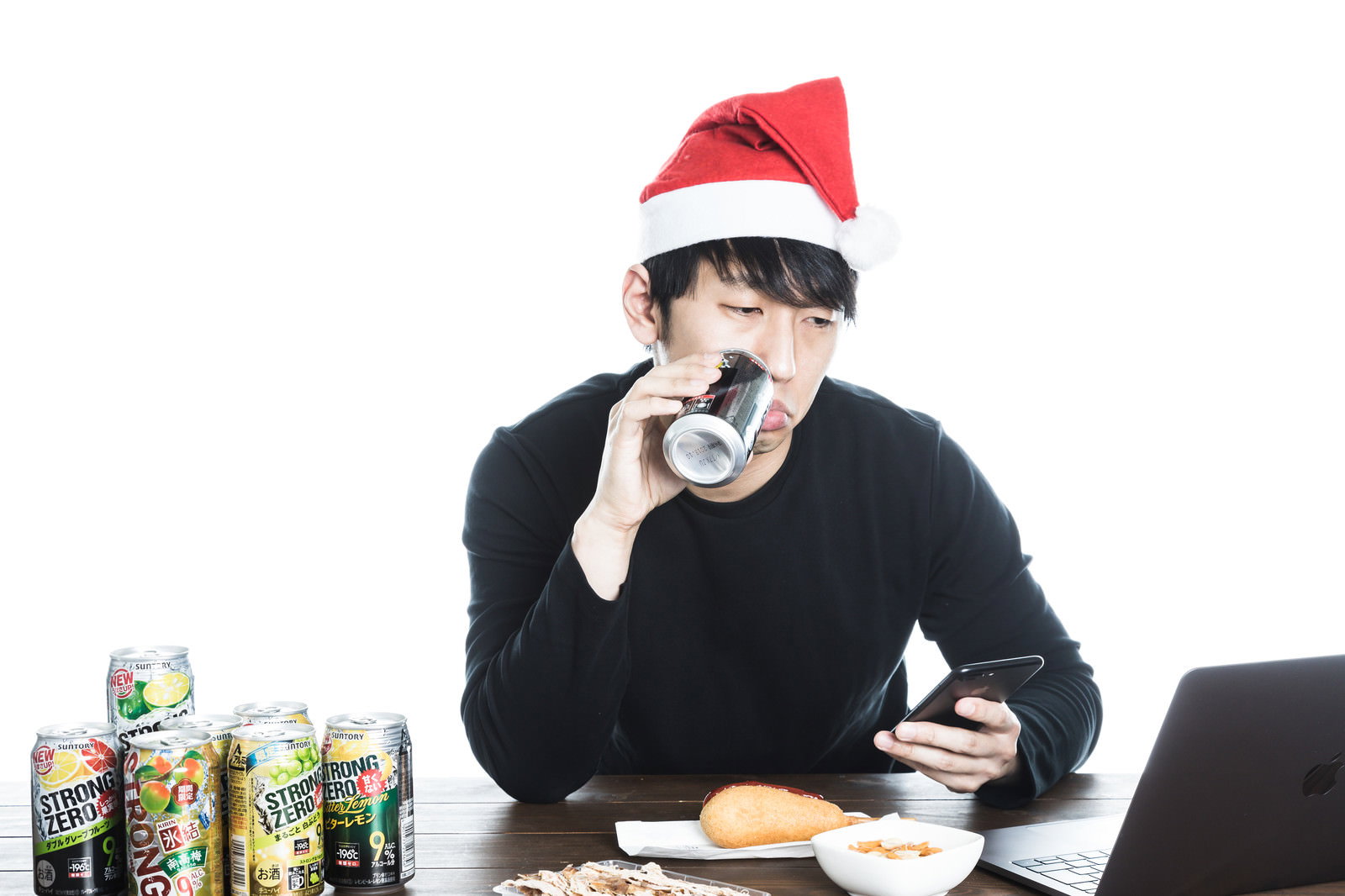 赤いサンタ帽をかぶった男性がテーブルで缶チューハイを飲みながらスマートフォンを操作している［モデル：大川竜弥］