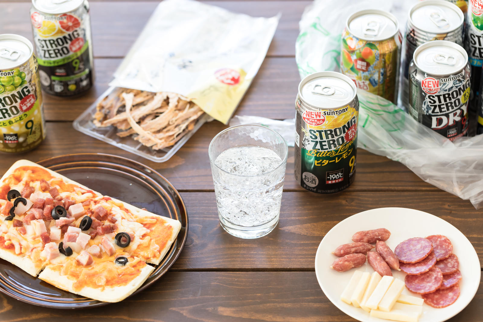 木製テーブルに置かれたピザやハムなどのおつまみとストロングゼロなどの缶チューハイ