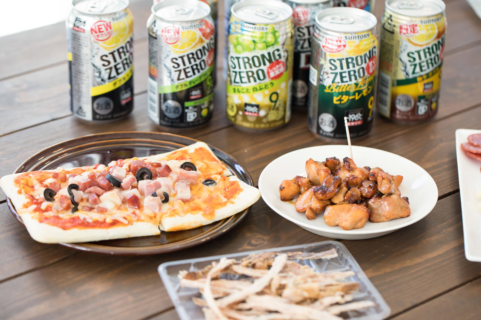木製テーブルにストロングゼロの複数缶、ピザ、唐揚げ、ナッツが配置された飲み会のシーン