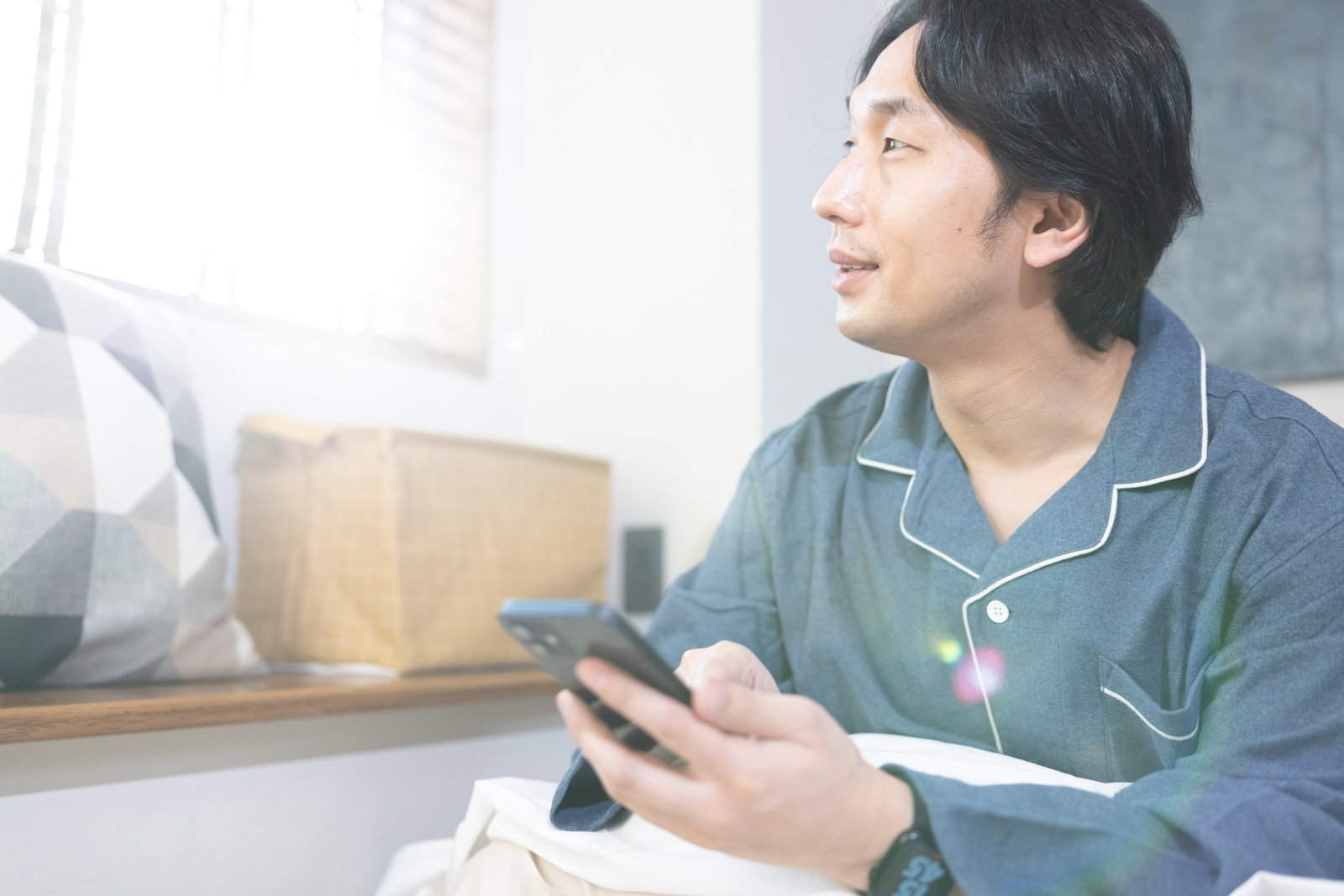 ベッドでスマートフォンを手に持ちながら朝の光の中で目覚める男性［モデル：大川竜弥］
