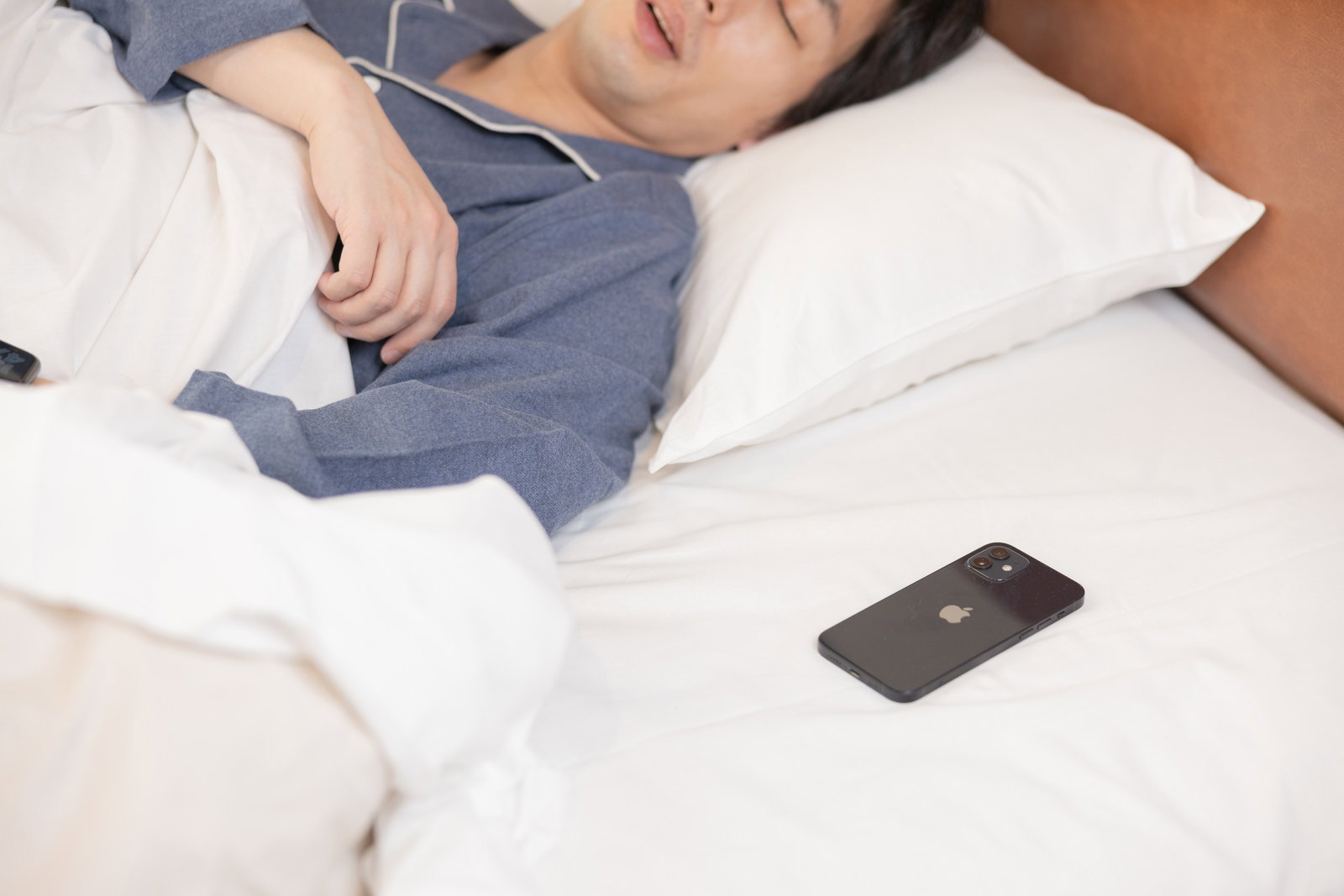 白いベッドで眠る男性の枕元に置かれたスマートフォン［モデル：大川竜弥］