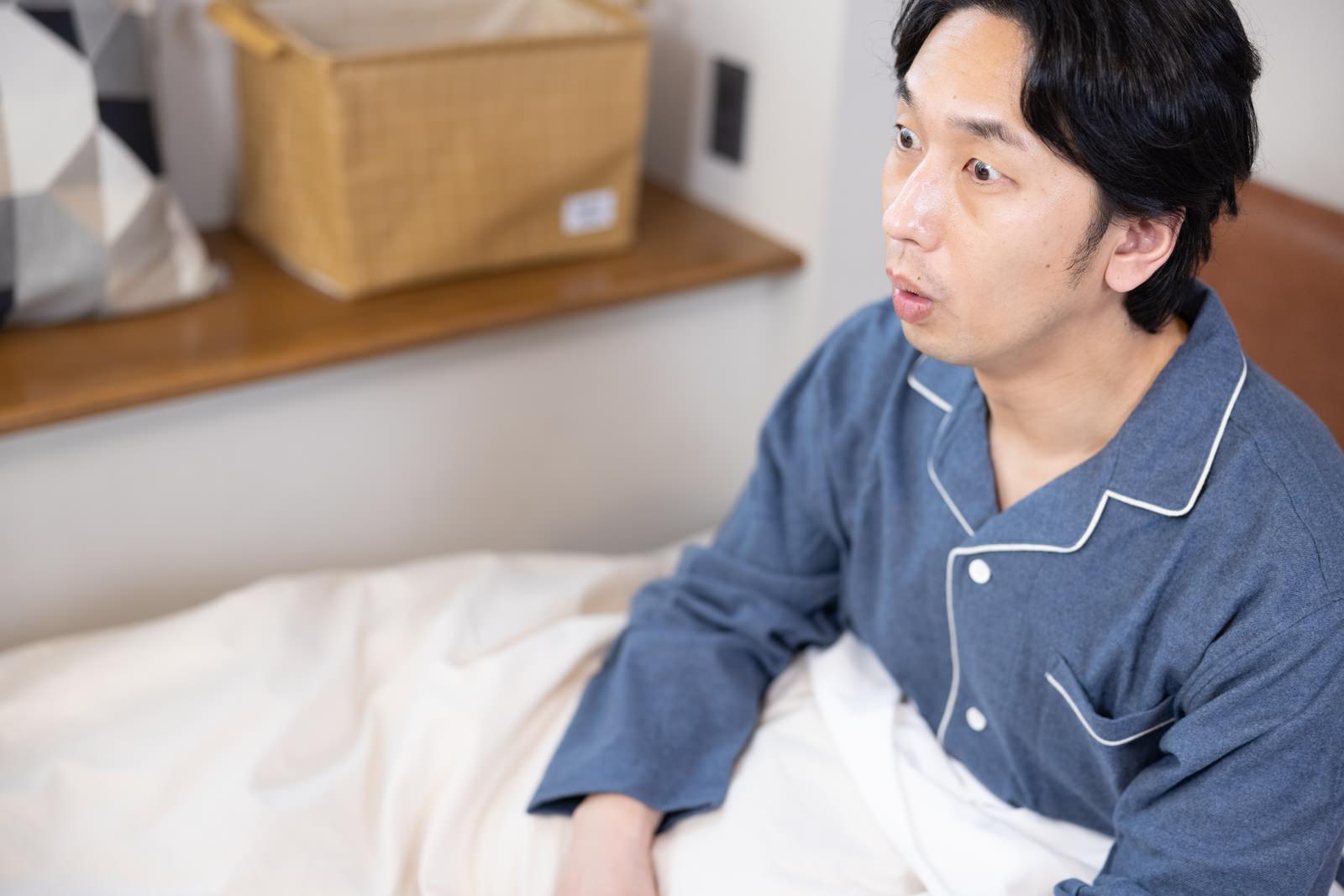 ベッドで目を覚まして時計を見て驚く表情のパジャマ姿の男性［モデル：大川竜弥］