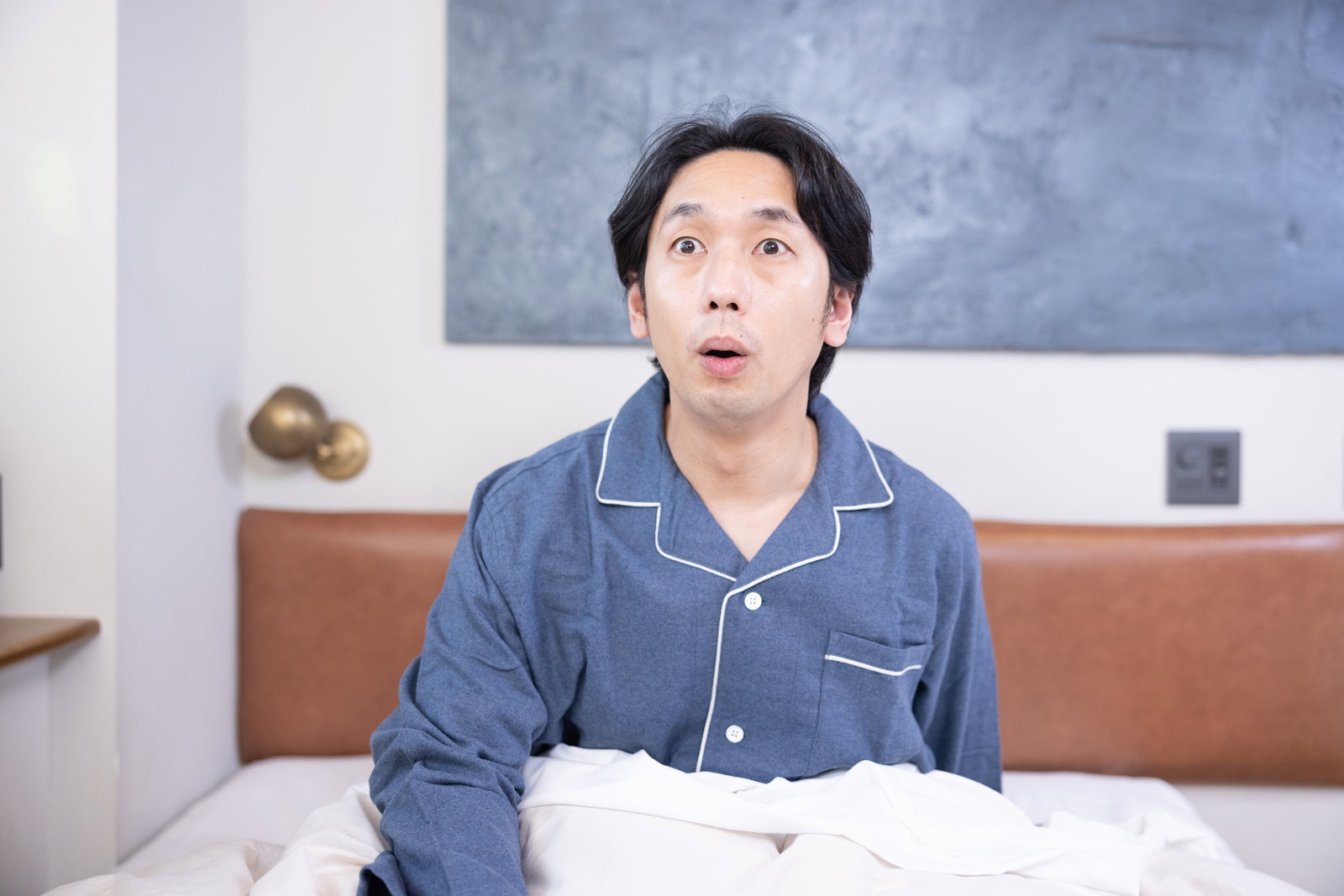 青いパジャマを着た男性がベッドの上で驚いた表情を見せている［モデル：大川竜弥］