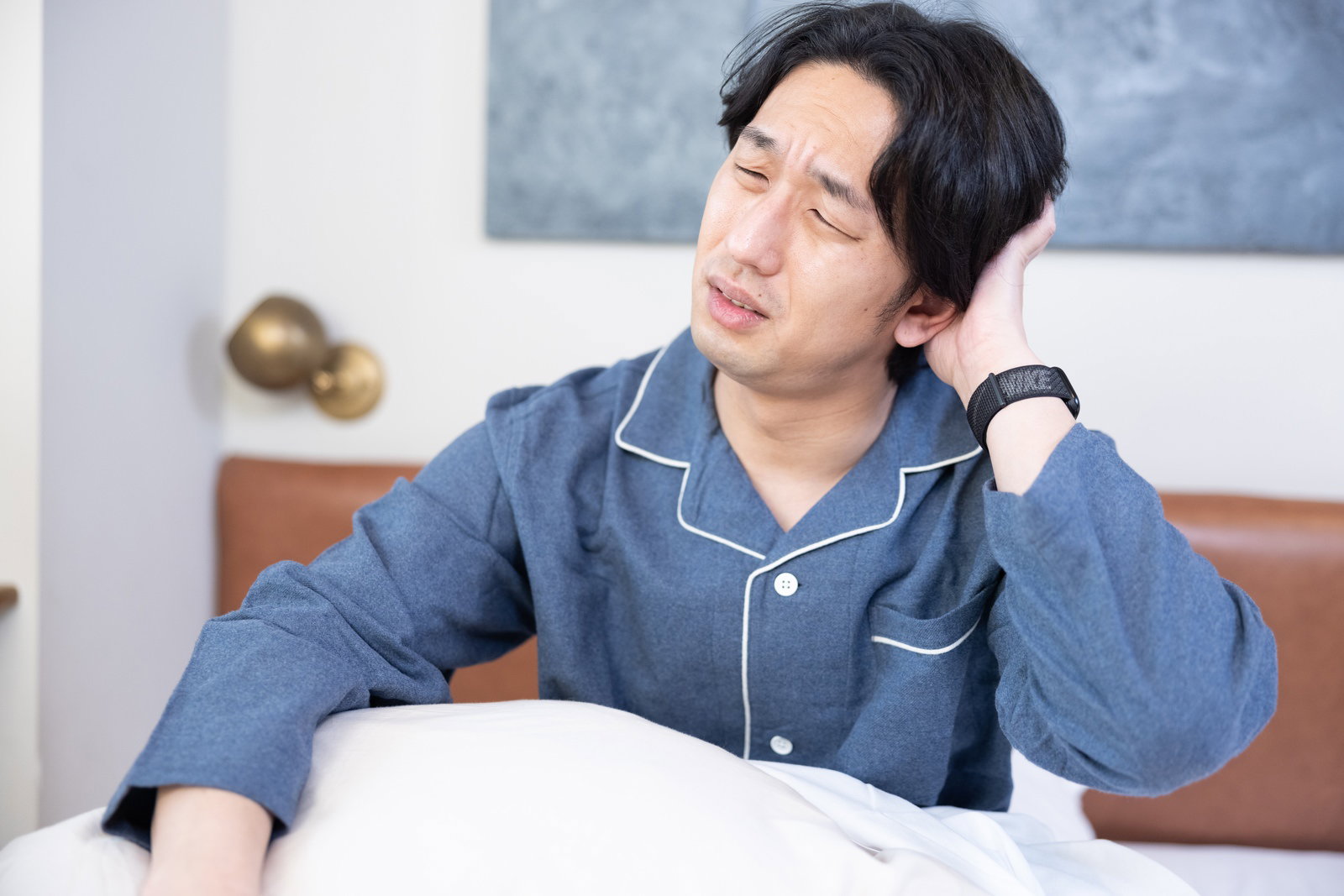 青いパジャマを着た男性がベッドの上で頭を抱えて困っている様子［モデル：大川竜弥］
