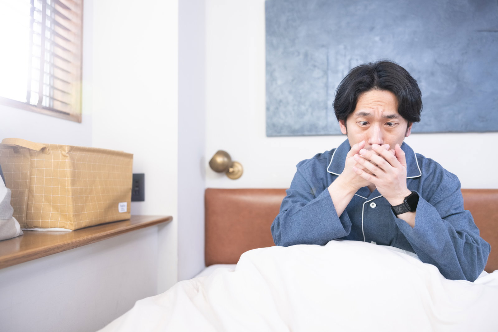 ベッドの上で咳をして手を口元に当てる男性［モデル：大川竜弥］