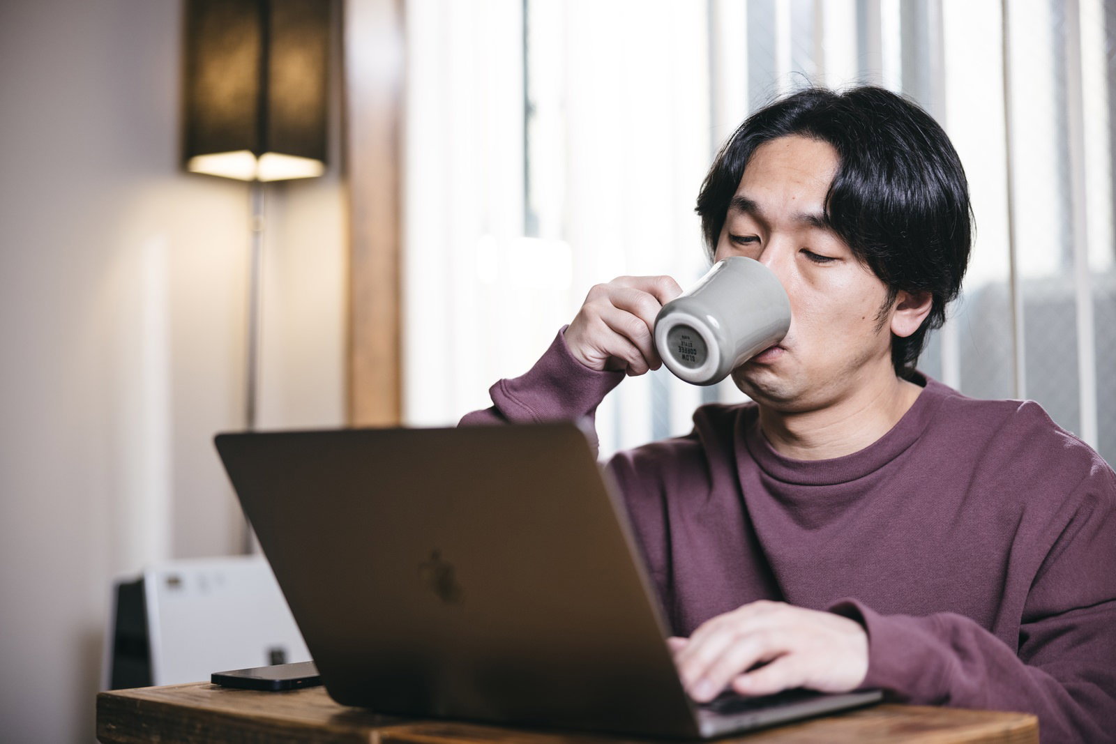 紫色のトップスを着た男性がノートパソコンの前でマグカップからコーヒーを飲んでいる様子［モデル：大川竜弥］