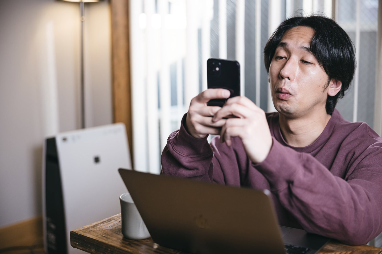 デスクでノートパソコンの前に座りスマートフォンを見る男性［モデル：大川竜弥］