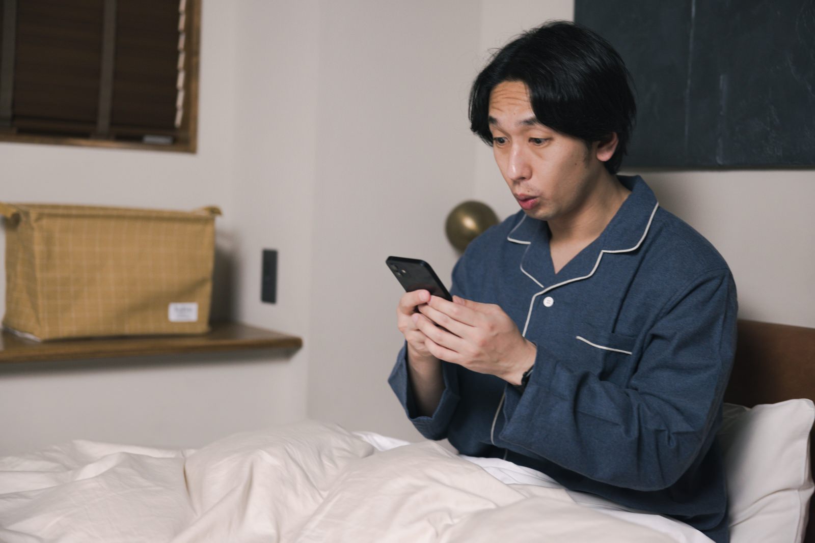 ベッドでスマートフォンを見て驚いた表情を浮かべる男性［モデル：大川竜弥］