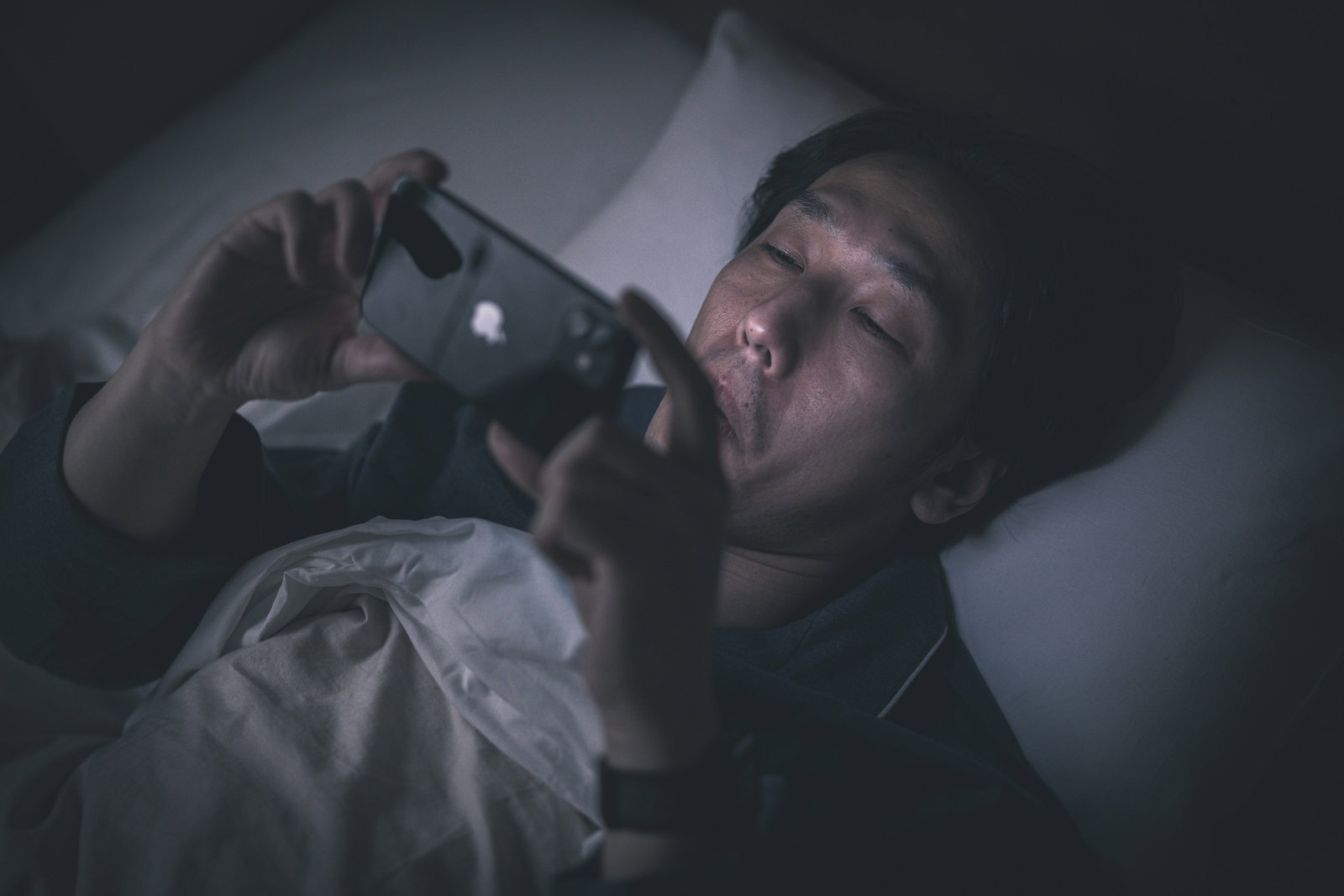 暗闇の中、ベッドでパジャマを着た男性がスマートフォンを見ている［モデル：大川竜弥］