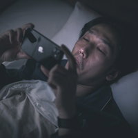 暗闇の中ベッドでスマートフォンを見る男性の写真