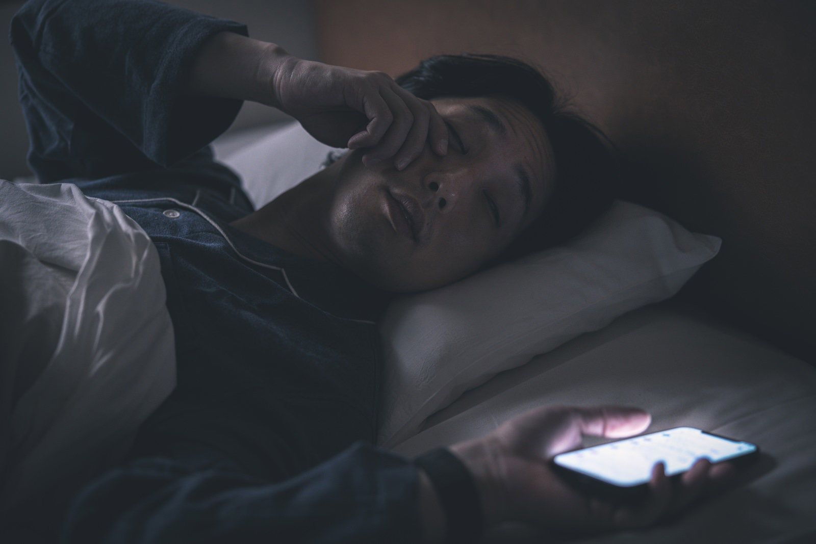 暗い部屋のベッドでスマートフォンを見ながら眠っている人［モデル：大川竜弥］