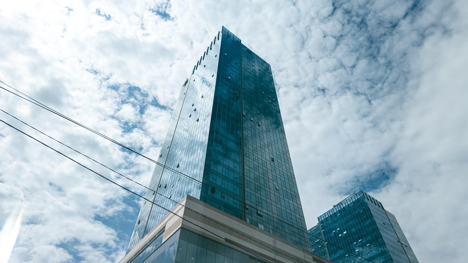 A glass-walled high-rise office building set against a blue sky and cloud background, shot from a low angle