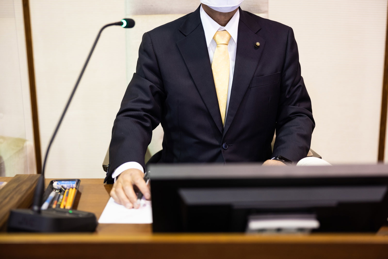 紺色スーツを着た議長が議会で書類をメモしながら前を見つめている