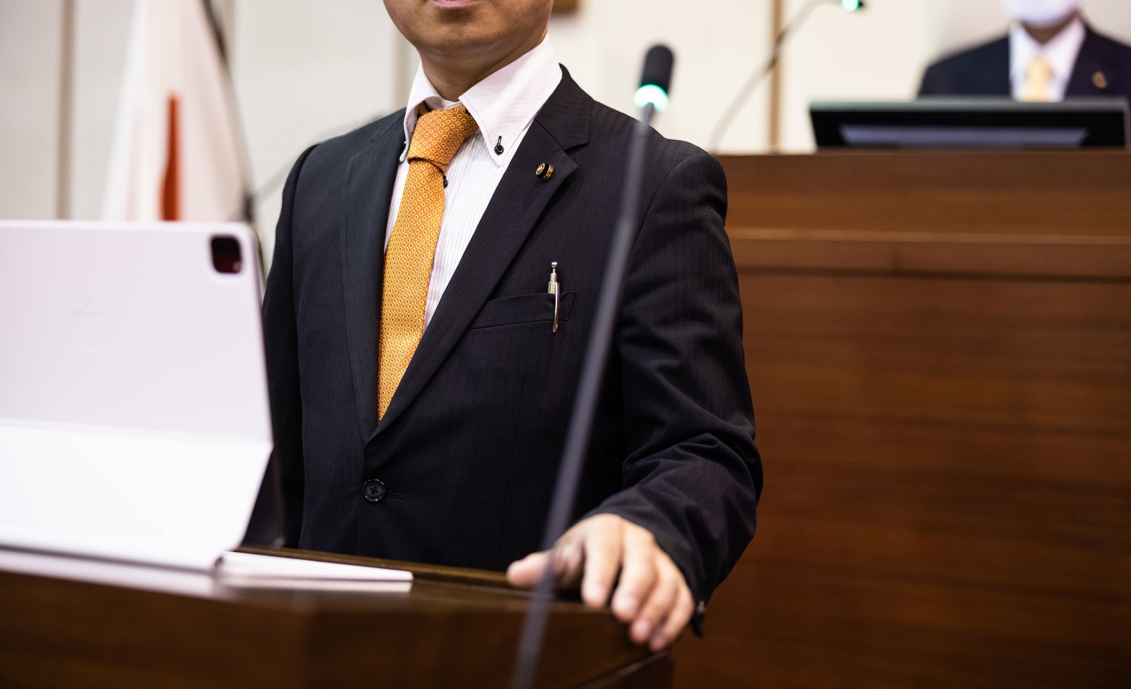 タブレット端末を持ち、議場で登壇する黒いスーツの男性議員