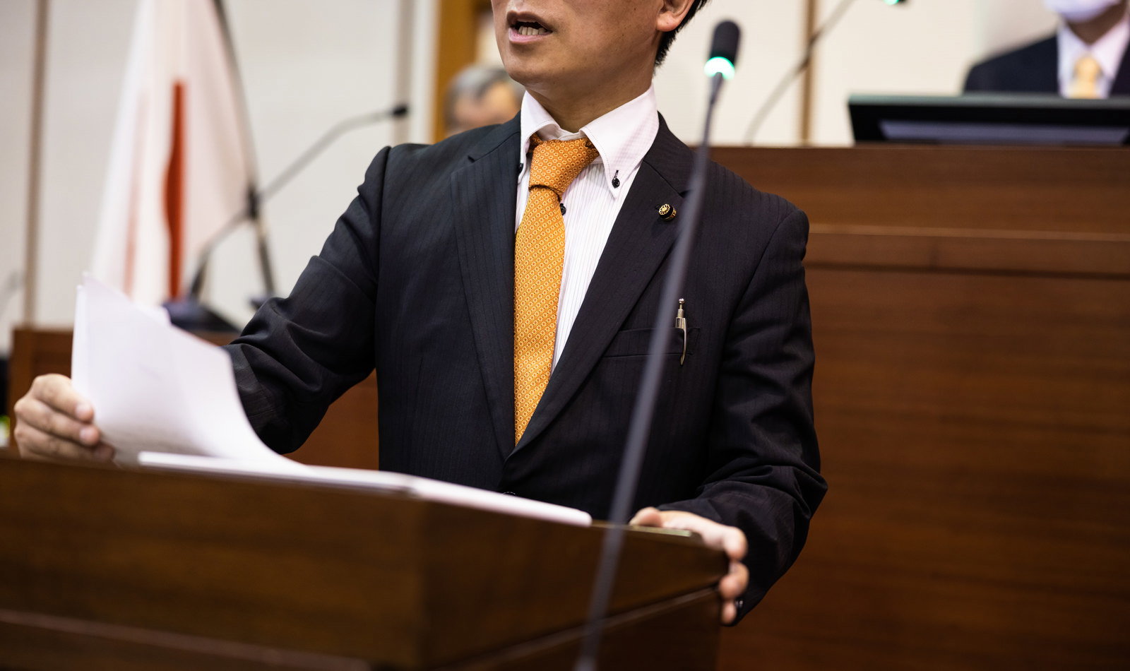 市議会の議場で金色ネクタイの議員が紙を手に登壇して発言している