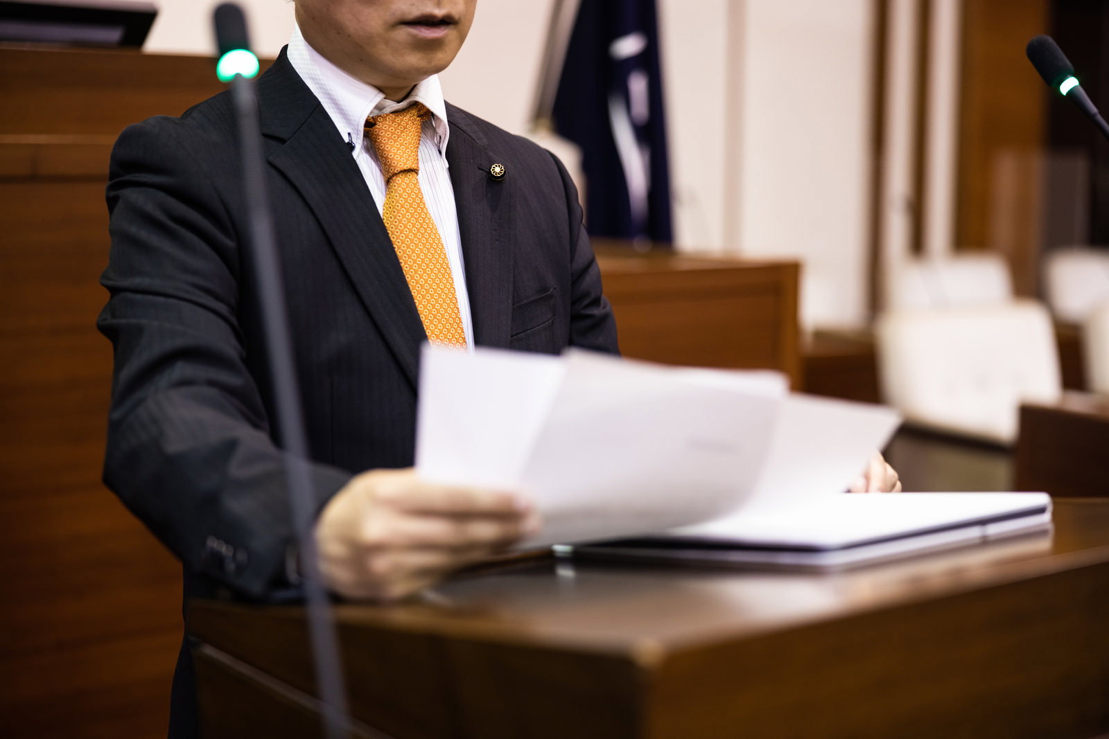黄橙色のネクタイをした市議会議員が原稿を持ちながら議場で発言している