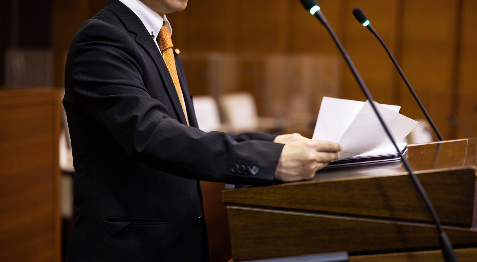 紺のスーツに金色ネクタイの議員が議会で原稿を読みながら演説している