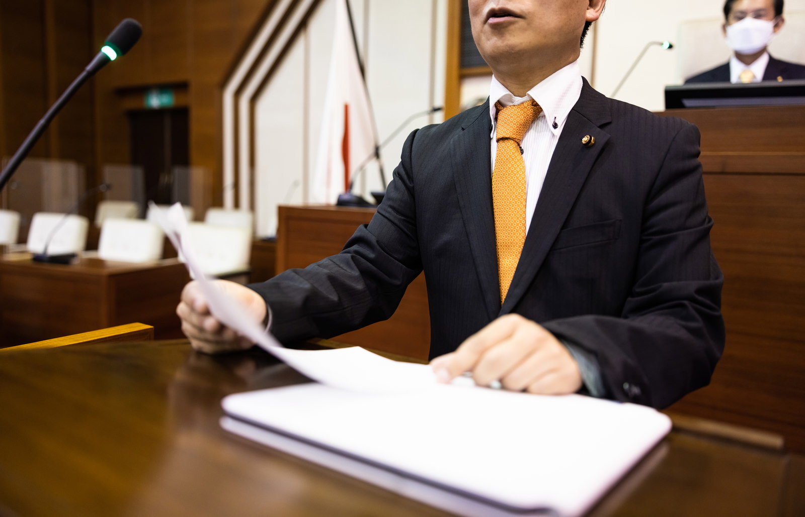 議場で発言する黒いスーツの議員と、背景の議長